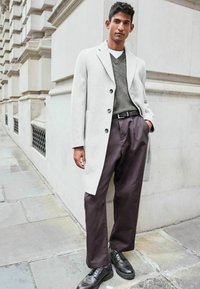 Abrigo gris claro sobre un suéter verde oscuro, combinado con pantalones oscuros sueltos y zapatos negros. Tela suave, corte a medida y detalles de botones.