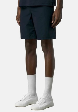 Uomo che indossa pantaloncini neri fino al ginocchio, calzini bianchi e scarpe da ginnastica bianche con lacci, in piedi davanti a uno sfondo semplice.