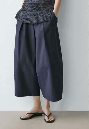 Persona che indossa pantaloni larghi e scampanati corti di colore scuro, una maglia senza maniche con trama e infradito nere, in piedi con le mani nelle tasche.