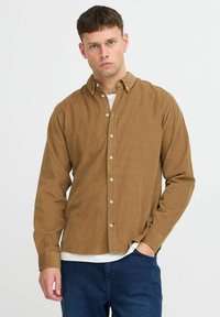 Brown corduroy shirt with button closure, long sleeves, and a relaxed fit. Worn over a white shirt, paired with dark blue trousers.