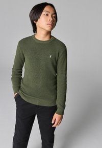 Maglione lavorato a maglia verde oliva con un motivo testurizzato, scollatura rotonda e polsini a coste, abbinato a pantaloni neri. Piccolo dettaglio del logo sul petto.