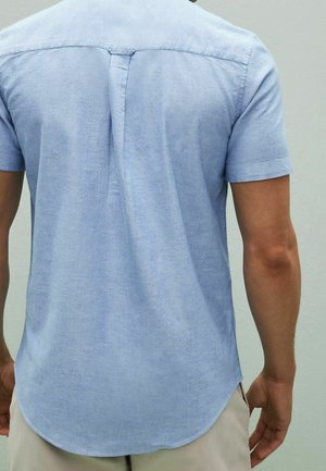 Krátký rukáv košile světle modré barvy, vyrobená z měkké látky s hladkým povrchem, s kulatým lemem a středním zadním skladem.