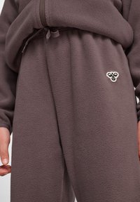 Fleece-Trainingsanzug in dunklem Taupe mit strukturiertem Stoff. Verfügt über einen Kordelzugbund und ein kleines gesticktes Bienenmotiv auf der Vorderseite.