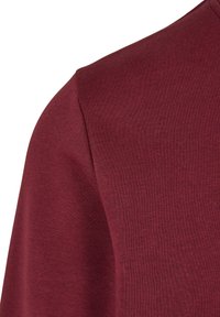 Burgundi színű pamuting sima tapintással, lekerekített nyakkivágással és rövid ujjal, kiemelve az anyag rugalmasságát és illeszkedését.