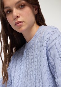 Jasnoniebieski sweter w splot kabli z teksturowymi wzorami i okrągłym dekoltem. Materiał wydaje się miękki, a krój jest luźny i wygodny.