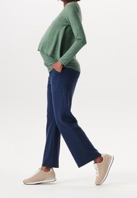 Kobieta w ciąży w zielonej bluzie z długim rękawem i granatowych spodniach, idąca z rękami w kieszeniach, w beżowych trampkach na białym tle.