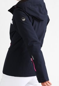 Voděodolná námořnicky modrá bunda s kapucí, zdobená růžovými zipy a logem na rukávu. Hladká, texturovaná látka.