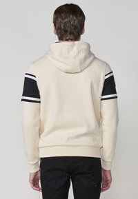 Hoodie v krémovej farbe s čiernymi a bielymi pruhmi na rukávoch. Obsahuje rebrované manžety a spodný lem, mäkkú textúru a voľný strih.