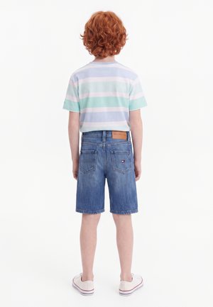 Barn med krøllet rødt hår står med ryggen til, iført lyseblå stribet t-shirt, denimshorts og hvide sneakers på en hvid baggrund.