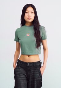 Green cropped t-shirt made of soft cotton, featuring a round neckline and a front logo design. Paired with dark, textured cargo pants.