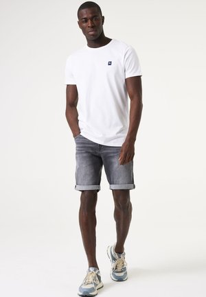Witte t-shirt met korte mouwen en een marineblauw logo, gecombineerd met grijze denimshorts met omgeslagen zoom. De schoenen hebben een gelaagd blauw en beige ontwerp.
