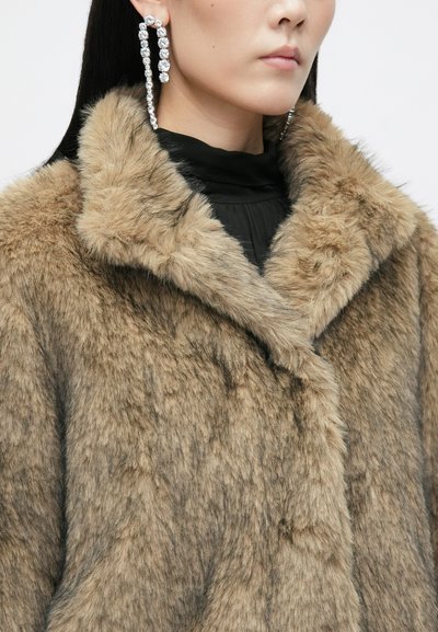 Μακριά καμπαρντίνα από τεχνητή γούνα σε ανοικτό καφέ χρώμα με μαλακή υφή, με φαρδύ γιακά και κλείσιμο με ένα κουμπί, φορεμένη πάνω από μαύρη μπλούζα.