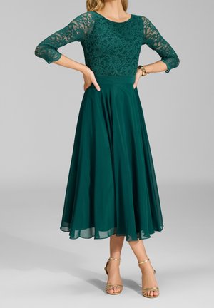 Femme portant une robe à dentelle vert foncé avec des manches trois-quarts et une jupe midi fluide, associée à des talons ouverts dorés.