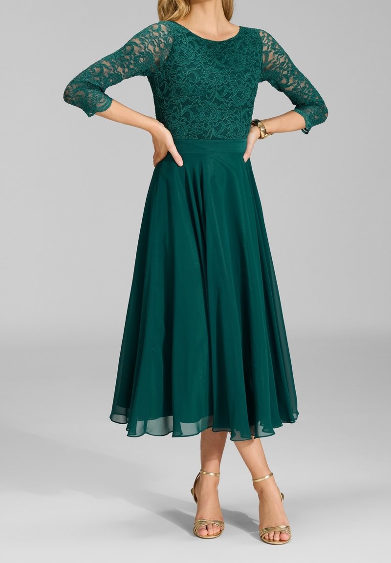 Femme portant une robe à dentelle vert foncé avec des manches trois-quarts et une jupe midi fluide, associée à des talons ouverts dorés.