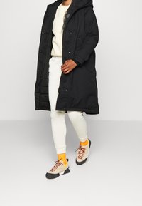 Černá dlouhá parka s kapucí, bílá mikina, světlé kalhoty a béžové tenisky s oranžovými akcenty a hnědými podrážkami.