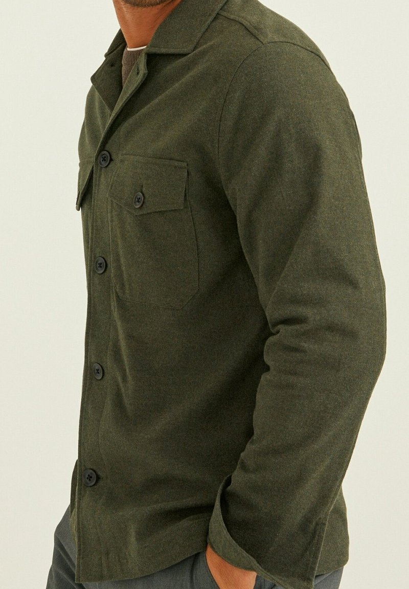 Homme portant une veste en laine vert olive boutonnée avec poche poitrine et manches longues sur fond uni.