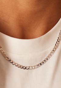 Zilveren ketting hangt op blote huid, rustend op een wit katoenen shirt. Bevat in elkaar grijpende schakels met een gladde, glanzende textuur.