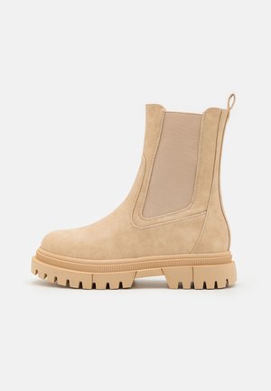 Botín Chelsea de ante beige con paneles elásticos en los laterales, suela de goma con tacos y un tirador para facilitar el calzado. Textura suave y líneas limpias.