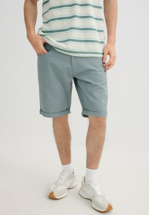 Hombre vestido con pantalones cortos azul claro arremangados, calcetines blancos, zapatillas blancas y una camisa de manga corta a rayas, de pie con las manos cerca de los bolsillos.