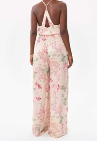 Kvinna iförd ärmlös, rygglös blommig jumpsuit med vida ben, ljusbeige bas, rosa och gröna blommönster samt guldarmband.