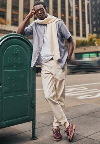 Model trägt ein hellgraues Kurzarmhemd, ein weißes T-Shirt, beige Hosen und kastanienbraune Schuhe und lehnt sich an einen grünen USPS-Briefkasten.