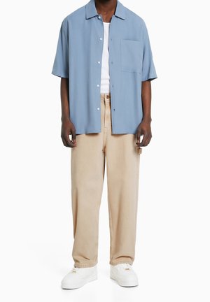 Hombre con camisa de manga corta color azul claro abotonada sobre camiseta blanca sin mangas, pantalones beige y zapatillas blancas, de pie contra un fondo blanco.