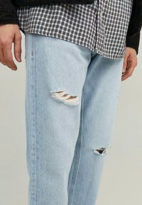 Jeans in denim azzurro chiaro con strappi usurati, caratterizzati da un fit rilassato e una texture sottile, abbinati a una camicia a scacchi bianca e nera.