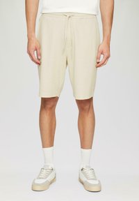 Pantalones cortos beige hechos de una tela suave y texturizada con una cinturilla elástica y cordón. Combinados con calcetines blancos y zapatillas beige.