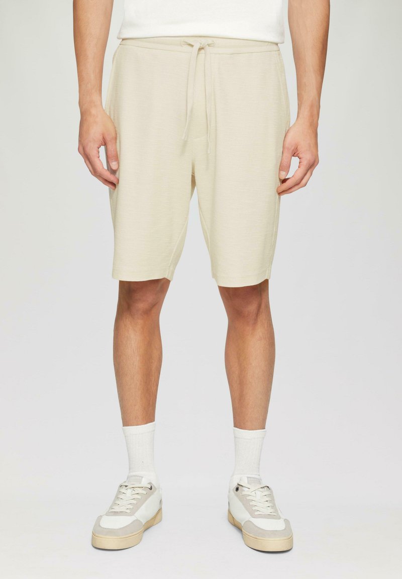 Pantalones cortos beige hechos de una tela suave y texturizada con una cinturilla elástica y cordón. Combinados con calcetines blancos y zapatillas beige.