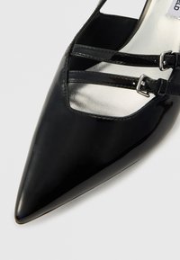 Μαύρη γόβα από λουστρίνι με μυτερή μύτη, με διπλές λουρίδες και ασημένια αγκράφια. Λείο υλικό με γυαλιστερό φινίρισμα.