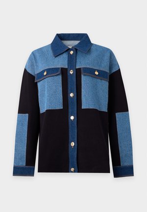 Femme aux longs cheveux bouclés portant une veste boutonnée bleue et noire, un t-shirt blanc, un jean large gris clair et des talons ouverts noirs.