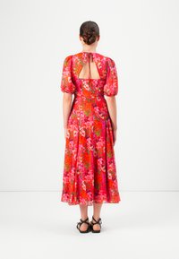 Woman with a low bun wearing a red floral midi dress with puff sleeves, open back detail, and black flat sandals, standing against a white backdrop.