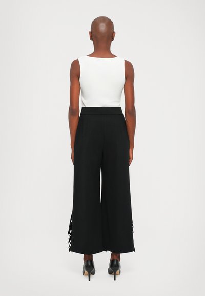 Top blanco sin mangas con escote redondo, combinado con pantalones negros de pierna ancha y cintura alta que presentan aberturas en el dobladillo. Zapatos de tacón negros.
