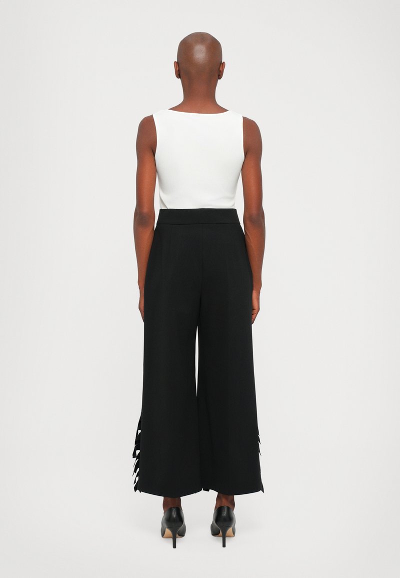 Witte mouwloze top met ronde hals, gecombineerd met hoge taille, wijde zwarte broeken met insneden aan de zoom. Zwarte hoge hakken.