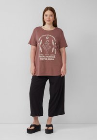 Braunes Oversized-T-Shirt mit weißem Grafikdruck, kombiniert mit schwarzen weiten Hosen und schwarzen Sandalen mit dickem Absatz. Einfaches Design und lässige Passform.