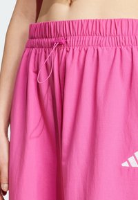 Růžové šortky s elastickým páskem, šňůrkou a bílým logem. Tkanina působí lehce a hladce, s jednoduchým designem.