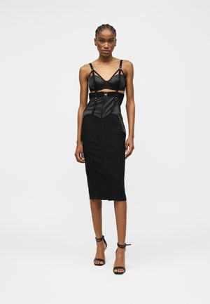 Žena v čiernych priliehavých midi šatách so strihaným pásom, štruktúrovaným živôtikom a tenkými ramienkami, doplnených čiernymi členkovými lodičkami.