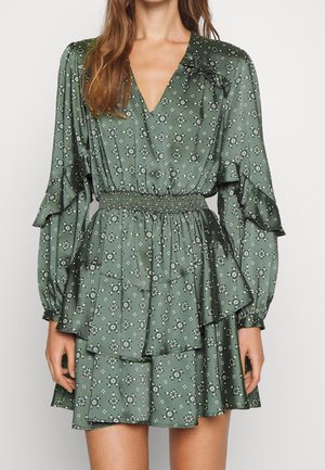 Vrouw draagt een groene jurk met patroon en lange mouwen, V-hals, gerimpelde lagen, elastische taille en gelaagde mouwen.