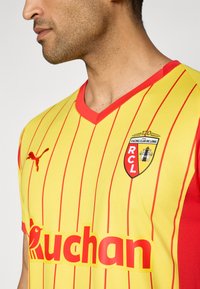 Un uomo che indossa una maglietta sportiva a righe gialle e rosse con il logo del Racing Club de Lens e la parola "Auchan" ben visibile.