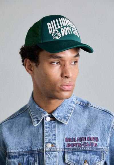 Casquette de camionneur verte avec le logo blanc "Billionaire Boys Club" et un graphique d'astronaute ; portée avec une veste en jean clair ornée de broderies rouges.