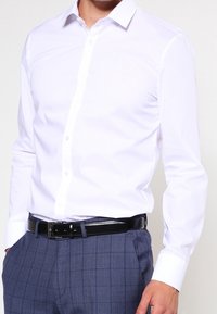 Camicia bianca con colletto, maniche lunghe e texture liscia, abbinata a pantaloni grigi a quadri e una cintura nera.