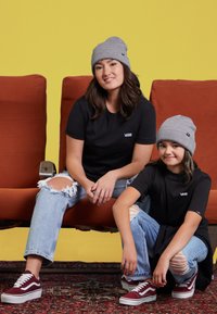 Svarta t-shirts med Vans-logga, ljusblå rivna jeans och gråa beanies. Sitter på en orange soffa mot en gul bakgrund.