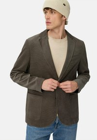 Grå kordfløjl blazer med to forlommer, notch-lapel og en let tekstureret overflade, båret over en beige skjorte og blå jeans.
