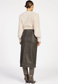 Vrouw die met haar rug naar de kijker staat, draagt een crèmekleurige gebreide trui met vlechtdetails, een donkergrijze leren midi-rok en een paar grijze hoge laarzen met hakken tot aan de knie.