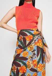 Top sans manches côtelé rouge avec col montant, associé à une jupe portefeuille à motif floral multicolore aux tons orange, vert et bleu.