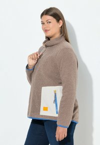 Brauner Fleece-Pullover mit hohem Kragen, ausgestattet mit einer kontrastierenden elfenbeinfarbenen Tasche und blauem Reißverschluss. Blaue Akzente am Saum und an den Ärmelbündchen verleihen dem Design das besondere Etwas.