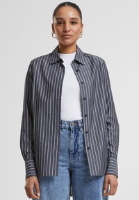 Donkergrijs button-up overhemd met witte verticale strepen, met een kraag, lange mouwen en een iets losse pasvorm. Draagt over een wit topje.