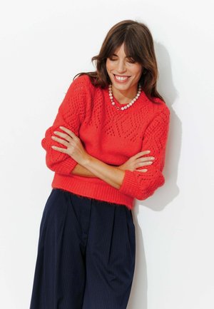Pull en maille épaisse rouge vif avec motif losange ; accompagné d'un pantalon plissé bleu marine. Accessoirisé d'un collier de perles.
