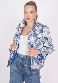 Floral-patterned jacket with blue, white, and green hues. Features a large collar and two front pockets. Worn with a white tank top.