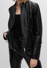 Kobieta ubrana w czarną skórzaną kurtkę z srebrnymi zamkami, nosząca czarną bluzkę oraz pasujące czarne skórzane spodnie, stoi z jedną ręką w kieszeni.